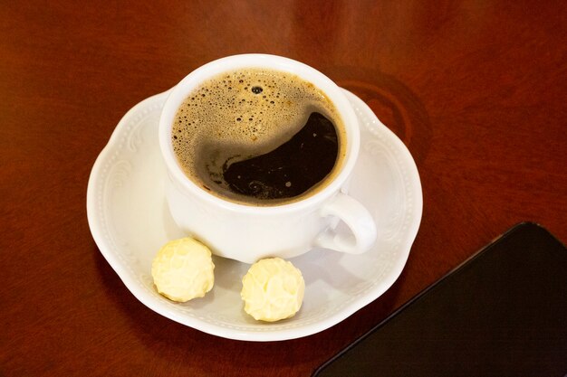 Uma xícara de café branco e balas redondas em um pires sobre uma mesa de madeira e um smartphone ao lado