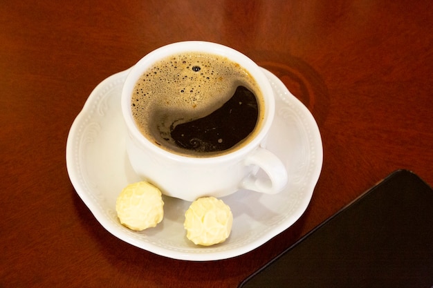 Uma xícara de café branco e balas redondas em um pires sobre uma mesa de madeira há um smartphone próximo