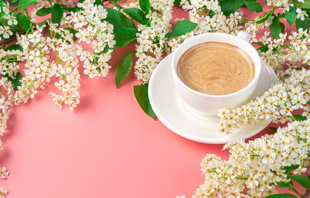Uma xícara de café branco em um pires entre flores em um fundo rosa. Vista lateral, copie o espaço.