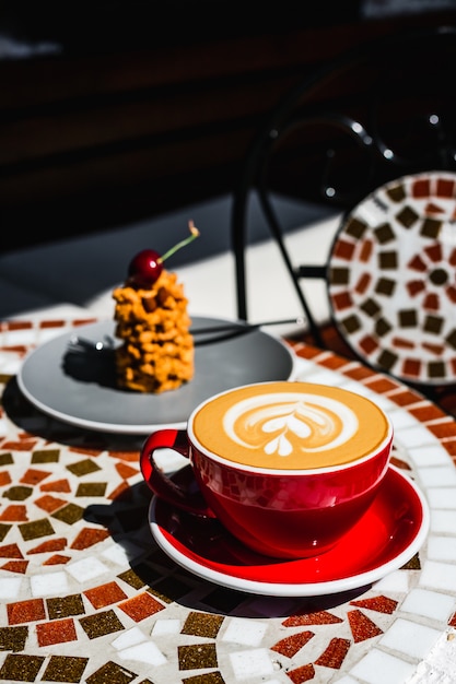 Uma xícara de café cappuccino vermelha feita na hora e uma sobremesa em uma mesa de pedra de mosaico de um terraço de café em um dia ensolarado