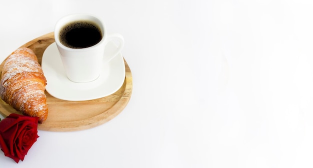 Uma xícara de café e um croissant em um suporte de madeira e uma rosa vermelha em um fundo branco