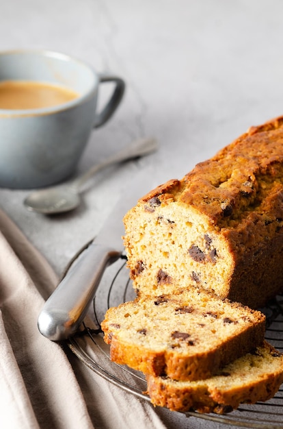 Uma xícara de café e um pedaço de pão de banana com gotas de chocolate em fundo cinza.