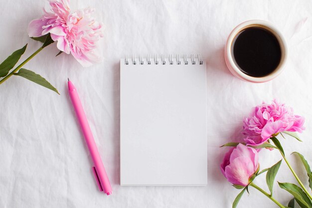 Uma xícara de café matinal no café da manhã, um bloco de notas vazio para seu texto ou desenho, flores de peônia rosa em uma toalha de mesa branca