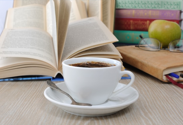 Uma xícara de café na mesa ao fundo de um livro aberto com um caderno e uma pilha de livros