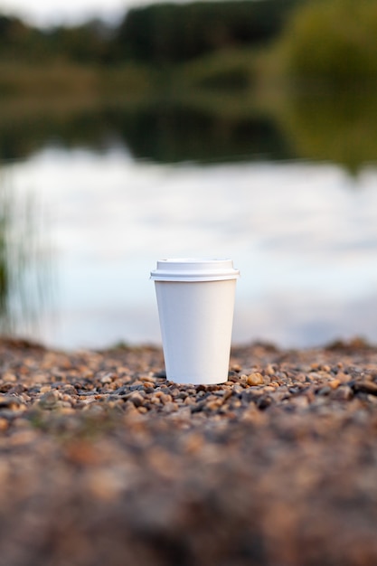 Uma xícara de café ou chá de papel branco fica na costa rochosa do lago. Uma caneca de bebida quente em uma pedra. Plano de fundo de beleza natural do lago na floresta atrás. espaço de cópia livre.