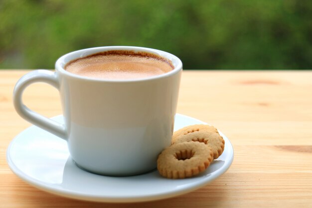 Uma xícara de café quente com biscoitos servidos na mesa de madeira com turva verde bush no fundo