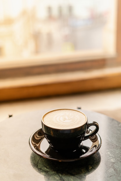 Uma xícara de cappuccino com espuma sobre uma mesa de mármore perto da janela em um pequeno e acolhedor café.