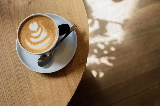 Uma xícara de cappuccino com latte art e colher em fundo de madeira. Espuma bonita, xícara de cerâmica branca.