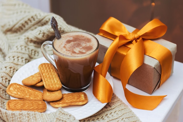 Uma xícara de chocolate e biscoitos em um prato branco sobre uma mesa branca e um presente com uma fita dourada