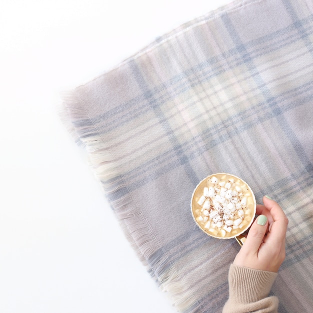 Uma xícara de chocolate e marshmallows em uma mão feminina em um lenço em pó quente