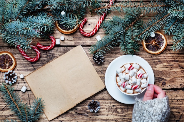 Uma xícara de chocolate quente com marshmallow nas mãos de um homem em um suéter. Árvore de Natal e decorações, caramelo de cana e nozes de laranjas Espaço em madeira da cópia do fundo. Presente de ano novo.