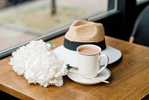 Uma xícara de chocolate quente perfumado com flores de hortênsia brancas e um chapéu na mesa de um café. Foco seletivo suave.