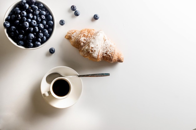 Uma xícara de croissant de café quente e mirtilos na mesa de tigela branca