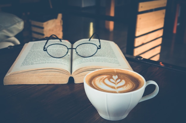 Uma xícara de latte art com óculos no livro aberto no café