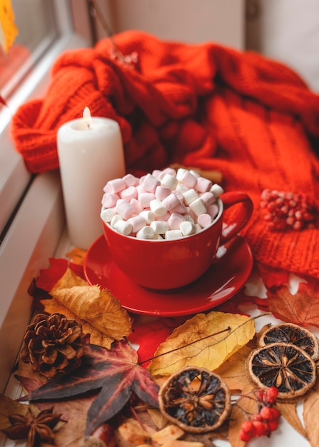 Uma xícara de marshmallow vermelho com um pires em folhas de outono com um lenço laranja uma vela um cone anice