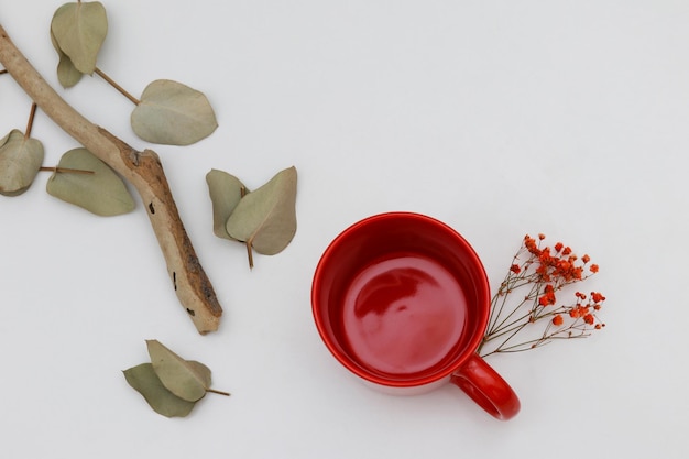 Uma xícara vermelha de café está sobre uma mesa branca ao lado de um galho com folhas e bagas.