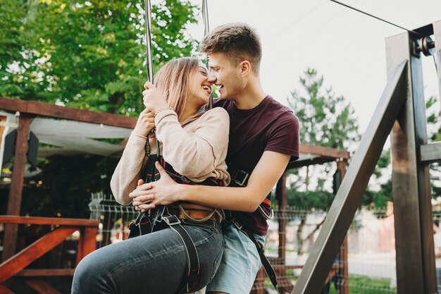 Umarmen des jungen Mannes und der jungen Frau im Sicherheitsgurt, der am Kabel der Seilrutsche hängt und vor der Fahrt mit Liebe lacht