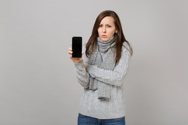 Umgekippte Frau im grauen Pullover, Schal, der Handy mit leerem leerem Bildschirm lokalisiert auf grauem Hintergrund hält. Gesunde Mode-Lifestyle-Leute aufrichtige Emotionen, Konzept der kalten Jahreszeit. Kopieren Sie Platz.