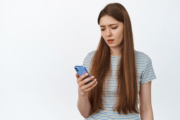 Umgekippte junge Frau, die ihr Handy anschaut, enttäuschte Smartphone-Nachricht liest und über weißem Hintergrund steht.