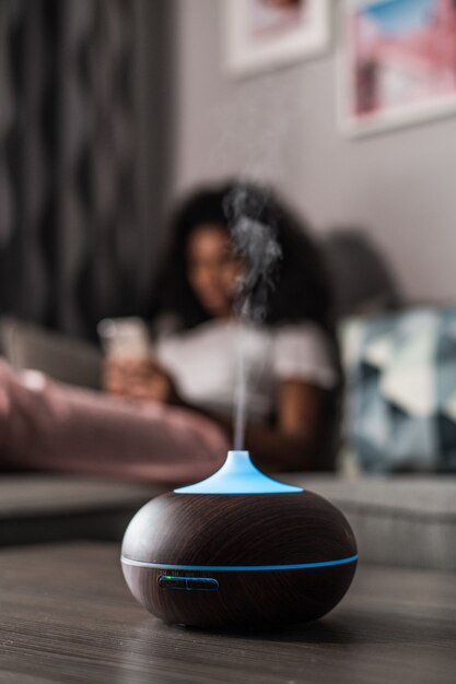 Foto umidificador de ar inteligente emitindo vapor d'água na mesa perto do sofá com uma mulher negra em casa