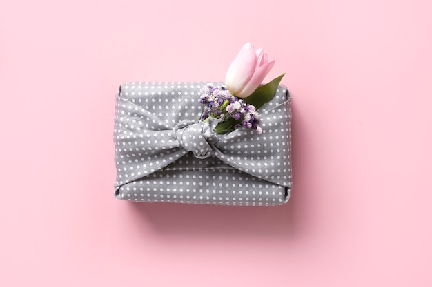 Umweltfreundliches Frühlingsgeschenk, eingewickelt in graues Textil mit Blumendekor auf Rosa. Nachhaltiges Geschenk. Kein Verlust. Osterferien. Traditioneller japanischer Furoshiki-Stil. Sicht von oben.
