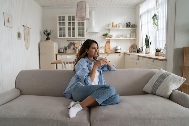 Unabhängige Frau, die mit einer Tasse Kaffee träumt, sitzt auf dem Sofa in einem geräumigen Haus und genießt die Einsamkeit
