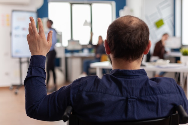 Unabhängiger behinderter Geschäftsmann, der ins Start-up-Büro kommt, winkt im Rollstuhl sitzend und grüßt das Geschäftsteam