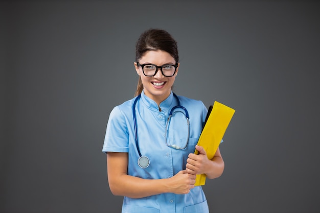 Unangenehme Einstellung einer Krankenschwester inter. Eine junge Krankenschwester in blauer Uniform mit Brille und Stethoskop um den Hals steht vor einer grauen Wand und hält einen Ordner mit Dokumenten