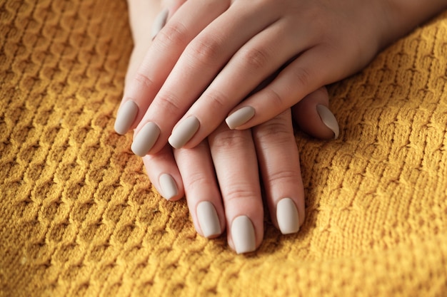 Uñas beige nude mate de cerca. Manicura de invierno u otoño, mano de mujer en suéter amarillo cálido