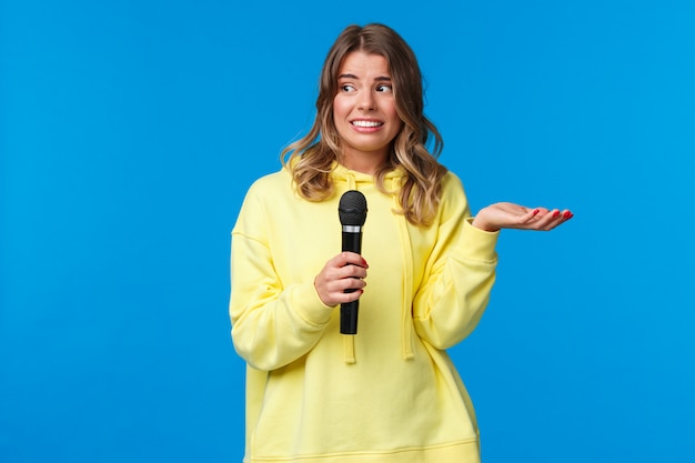 Unbeholfenes süßes blondes Mädchen kennt keine Texte als Karaoke-Gesang, Blick auf Freund nervöses Lächeln, verlegen singen vor Menschen, Mikrofon halten, blaue Wand stehen