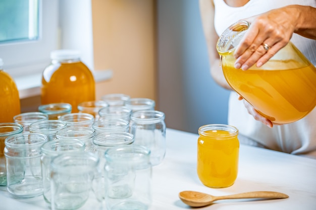Unbekannte Frau gießt süßen goldenen Honig in transparente, leere, saubere Gläser, die an einem hellen, sonnigen Tag auf einem weißen großen Holztisch stehen