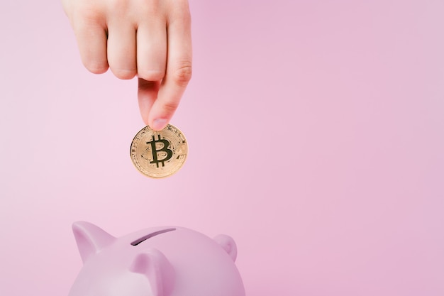 Unbekannte Hand mit einem silbernen Bitcoin und einem rosa Sparschwein auf einem Hintergrund, der in Kryptowährung spart