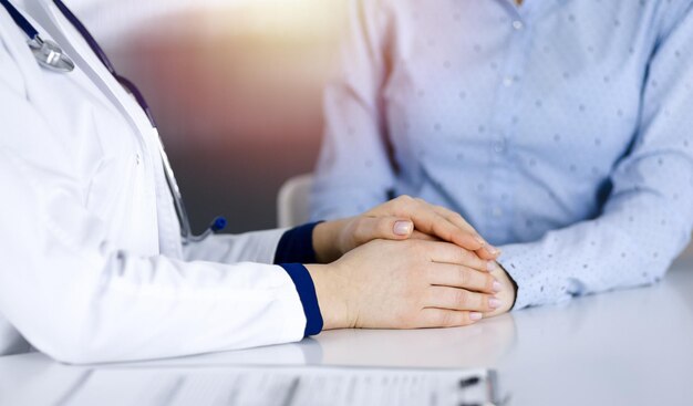 Unbekannte Ärztin hält die Hände ihrer Patientin, um eine Patientin zu beruhigen, während sie gemeinsam am Schreibtisch im sonnigen Kabinett einer Klinik sitzen. Ärztin benutzt ein Klemmbrett und ein Stethoskop,