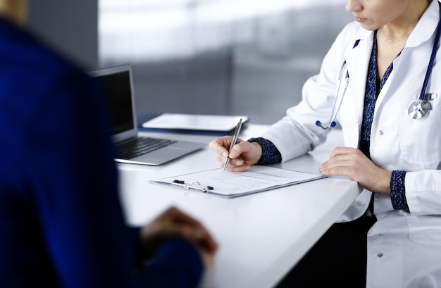 Unbekannte Ärztin schreibt ihrer Patientin medizinische Empfehlungen, während sie gemeinsam am Schreibtisch im Schrank einer Klinik sitzen. Arzt benutzt ein Klemmbrett, Nahaufnahme. Perfekt m