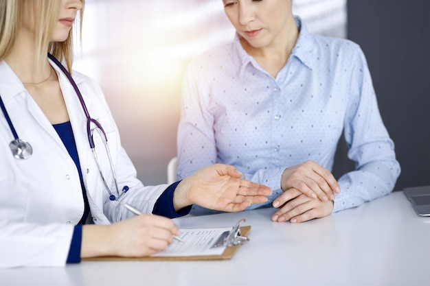 Unbekannte Ärztin und ihre Patientin schauen sich die aktuellen medizinischen Testergebnisse der Patientin an, während sie gemeinsam am Schreibtisch im sonnigen Schrank einer Klinik sitzen. Ärztin benutzt ein Klemmbrett