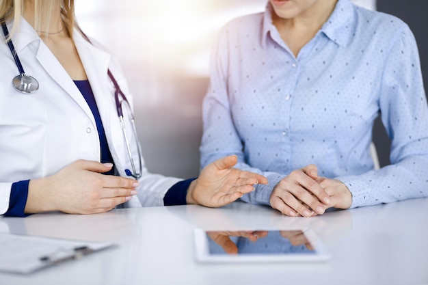 Unbekannte Ärztin zeigt ihrer Patientin eine Medikamentenbeschreibung, während sie gemeinsam am Schreibtisch im sonnigen Schrank einer Klinik sitzen. Ärztin benutzt ein Computertablett und einen Steth