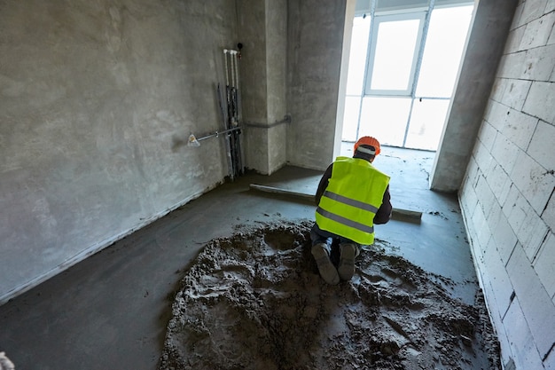 Foto unbekannter arbeiter in schutzkleidung und orangefarbenem schutzhelm macht in einer wohnung eines im bau befindlichen hochhauses einen zementestrich