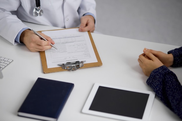 Unbekannter bärtiger Arzt und Patientin diskutieren aktuelle Gesundheitsuntersuchung, während sie in sonniger Klinik sitzen, Nahaufnahme. Medizin-Konzept.