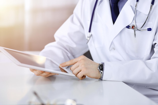 Unbekannter männlicher Arzt sitzt und arbeitet mit einem Tablet-Computer in einer abgedunkelten Klinik, grelles Licht im Hintergrund, Nahaufnahme von Händen