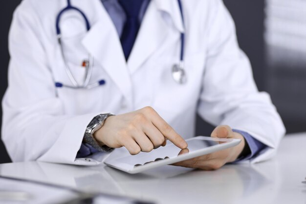 Unbekannter männlicher Arzt sitzt und arbeitet mit Tablet-Computer in der Klinik an seinem Arbeitsplatz, Nahaufnahme. Junger Arzt bei der Arbeit. Perfekter medizinischer Service, Medizinkonzept.
