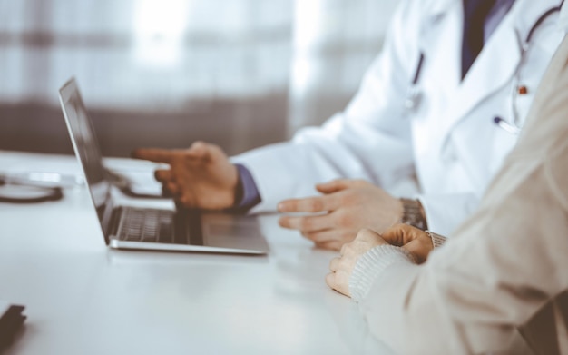 Unbekannter männlicher Arzt und Patientin diskutieren etwas, während sie in der Klinik sitzen und einen Laptop benutzen. Bester medizinischer Service im Krankenhaus, Medizin, Pandemiestopp.