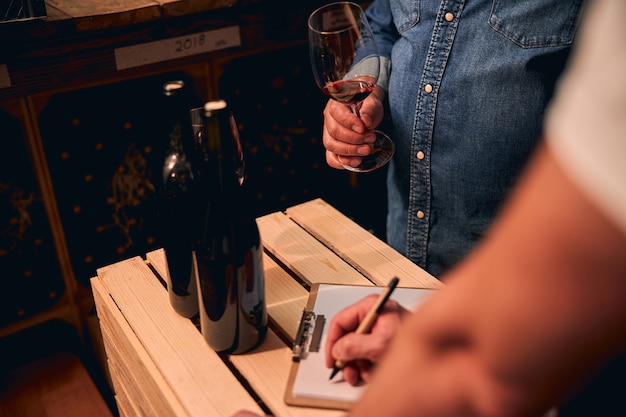 Unbekannter männlicher Sommelier, der mit einem Glas Rotwein im Keller steht, während sein Assistent sich Notizen macht