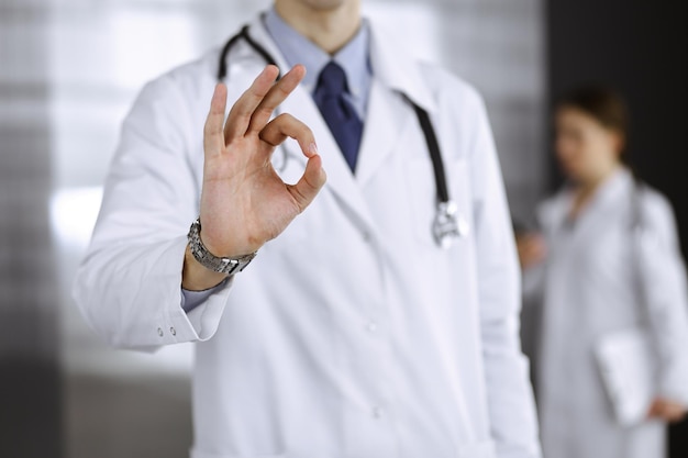 Unbekannter Mann-Arzt, der gerade steht und in der modernen Klinik ein Ok-Zeichen zeigt. Kollegin hat es eilig im Hintergrund des Arztes. Medizin-Konzept.