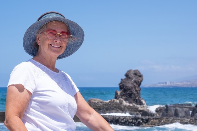 Unbeschwerte Seniorin, die am Strand sitzt und die Sommerferien genießt, blaue Mütze und rosa Sonnenbrille trägt. Entspannter Rentner, der in die Kamera schaut