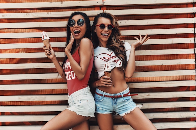 Unbeschwerte Zeit genießen. Zwei attraktive junge Frauen lächeln und halten Eis, während sie draußen an der Holzwand stehen