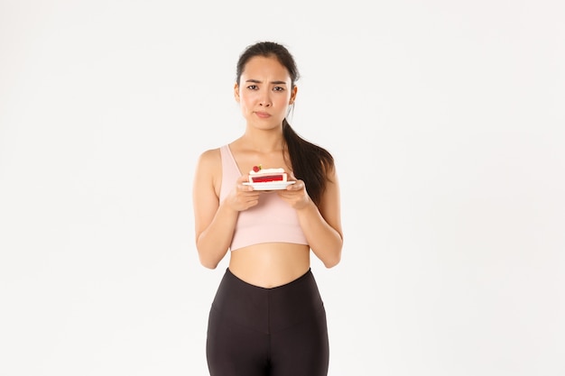 Unentschlossene niedliche asiatische Sportlerin mit Kuchen, der beunruhigt aussieht, will Süßigkeiten essen, an Kalorien und Körpergewicht nach dem Training denkend, weißer Hintergrund.