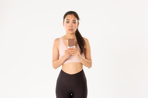 Unentschlossenes süßes asiatisches Mädchen in Trainingskleidung, das zögernd auf Schokoriegel schaut und versucht, Versuchung zu widerstehen, Gewicht auf Diät zu verlieren.