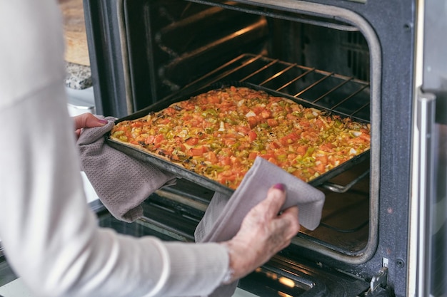 Unerkennbare ältere Frau beim Kochen, die Gemüse-Pizza auf dem Backbrett in den geöffneten Ofen in der Küche legt
