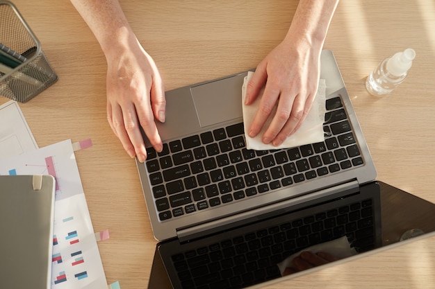 unerkennbare Frau, die Laptop während der Arbeit am Schreibtisch im Post-Pandemie-Büro desinfiziert, das durch Sonnenlicht beleuchtet wird