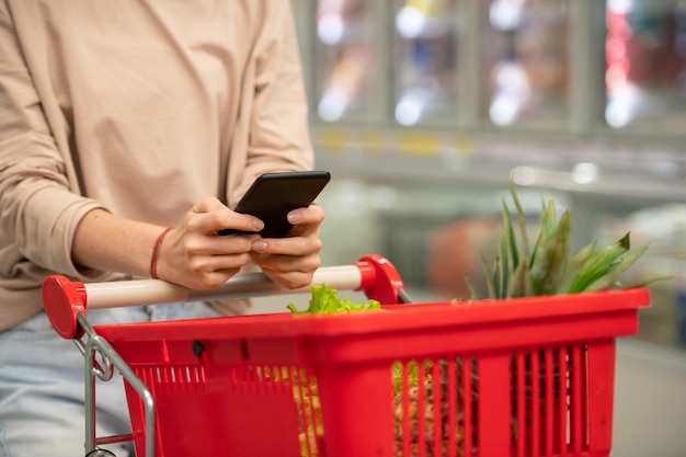 Unerkennbare Frau mit Einkaufswagen im Supermarkt, die die Einkaufsliste auf dem Smartphone überprüft